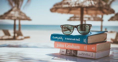 Et stabel med bøker av John Grisham og Cecelia Ahern med et par solbriller plassert oppå, ligger på en trebordkant med en strand og parasoller i bakgrunnen, som formidler en avslappende feriefølelse med sommerlesning og feriedager.